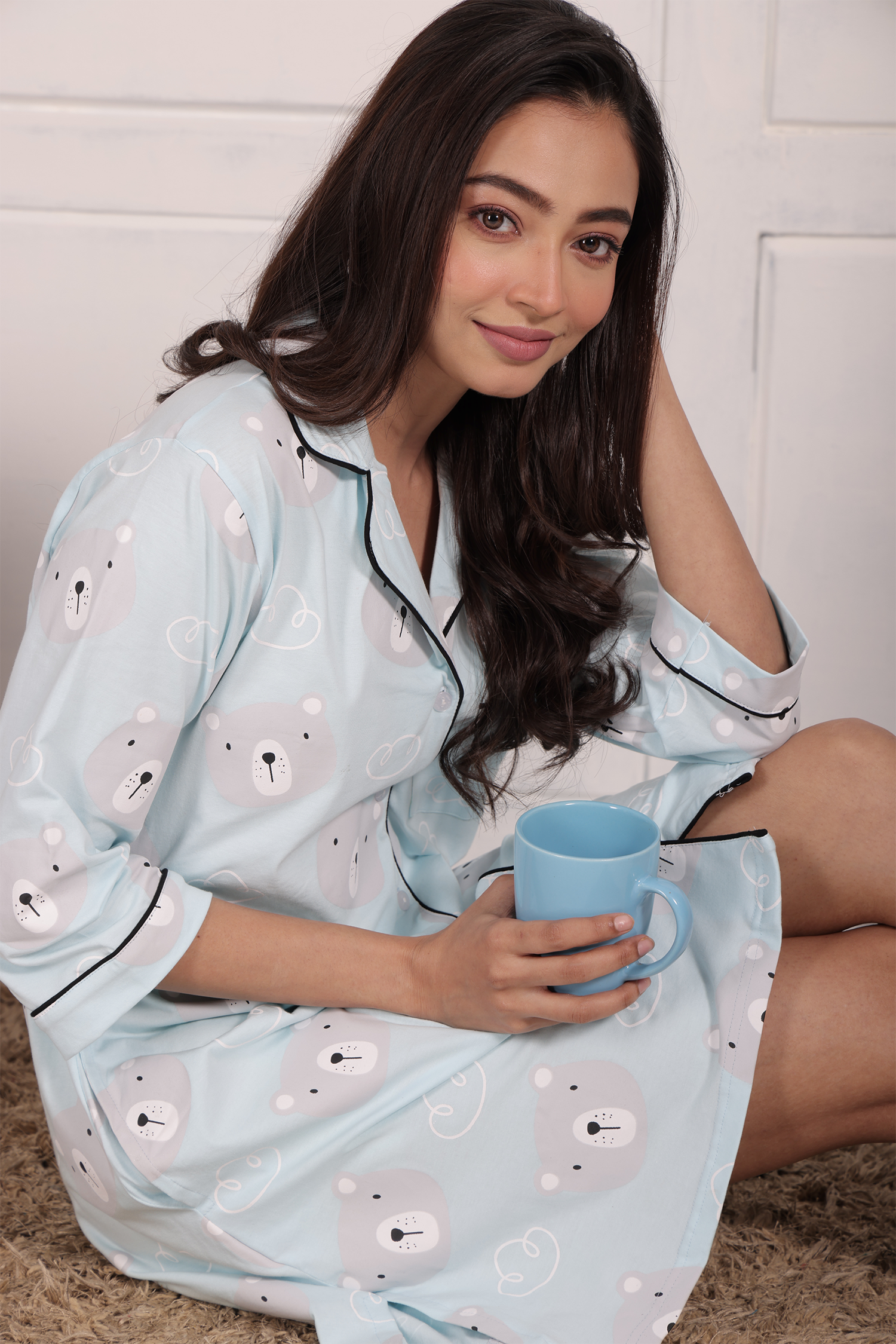 A girl sitting in style, holding a cup of coffee, and wearing a Teddy Print Sleep Shirt nightwear by NapStory.