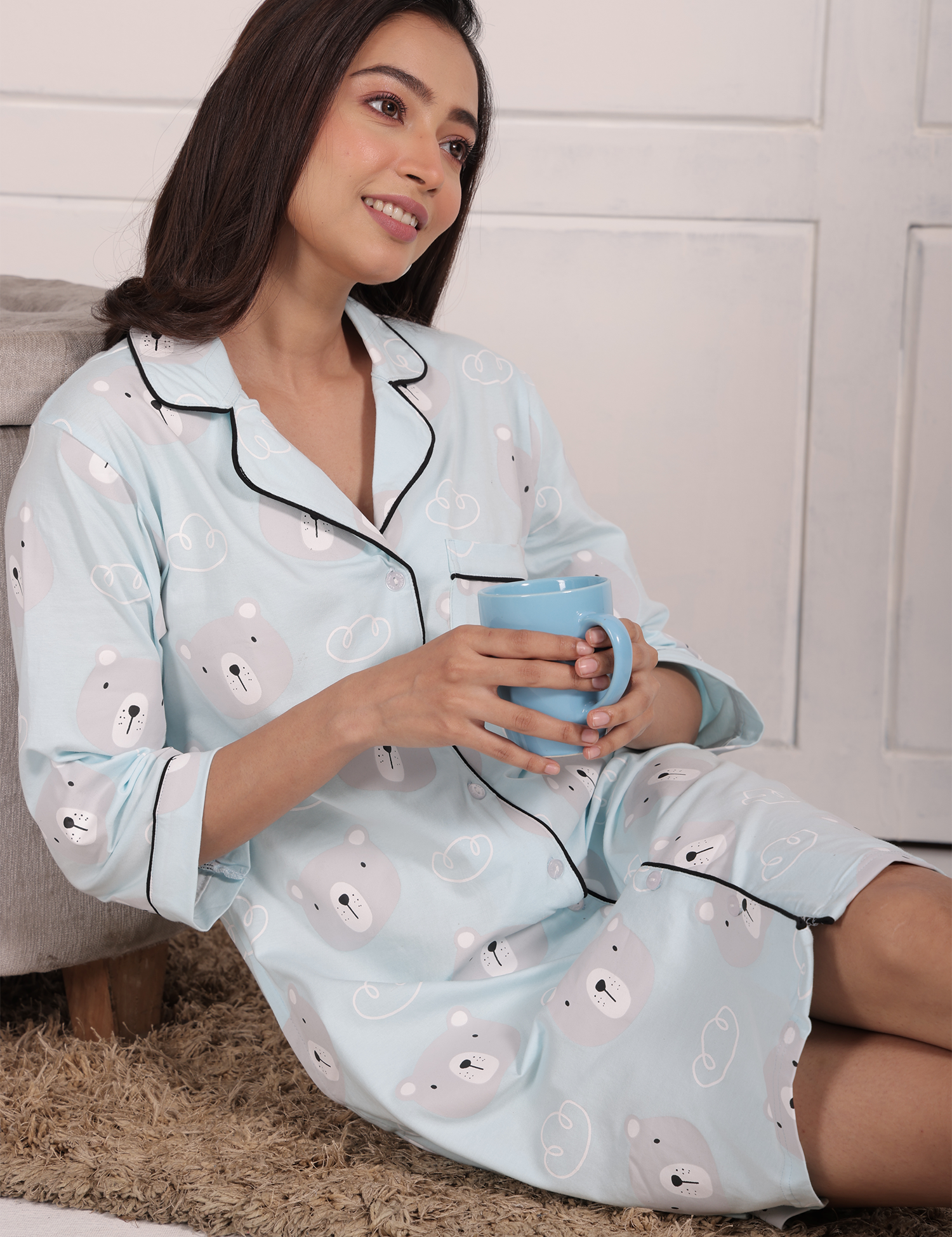 A girl reclining, holding a cup with both hands, wearing a Teddy Print Sleep Shirt nightwear by NapStory