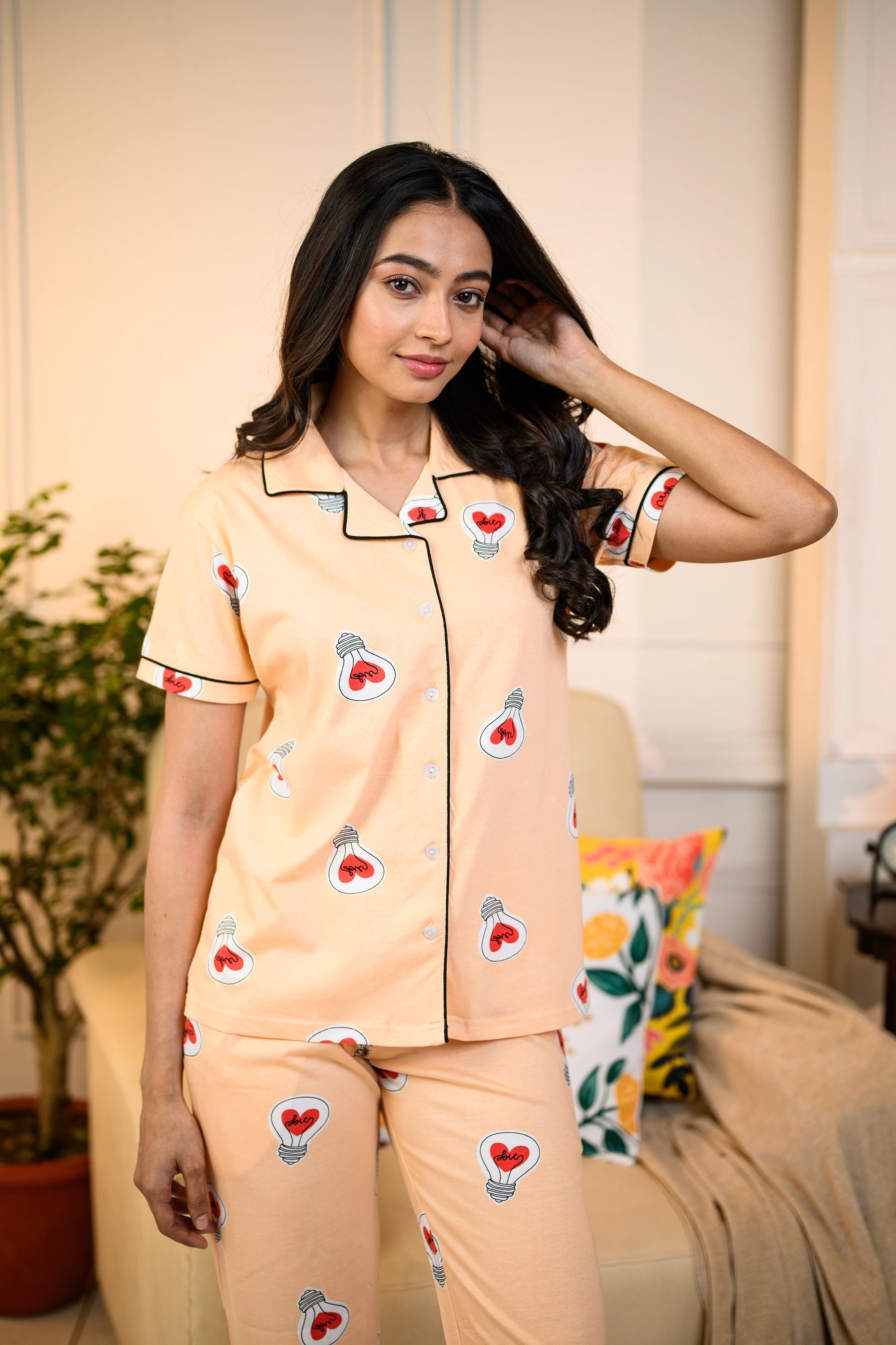 Woman in a peach Love Bulb Print Pyjama Set by NapStory, featuring playful bulb designs, posing casually in a cozy living space.