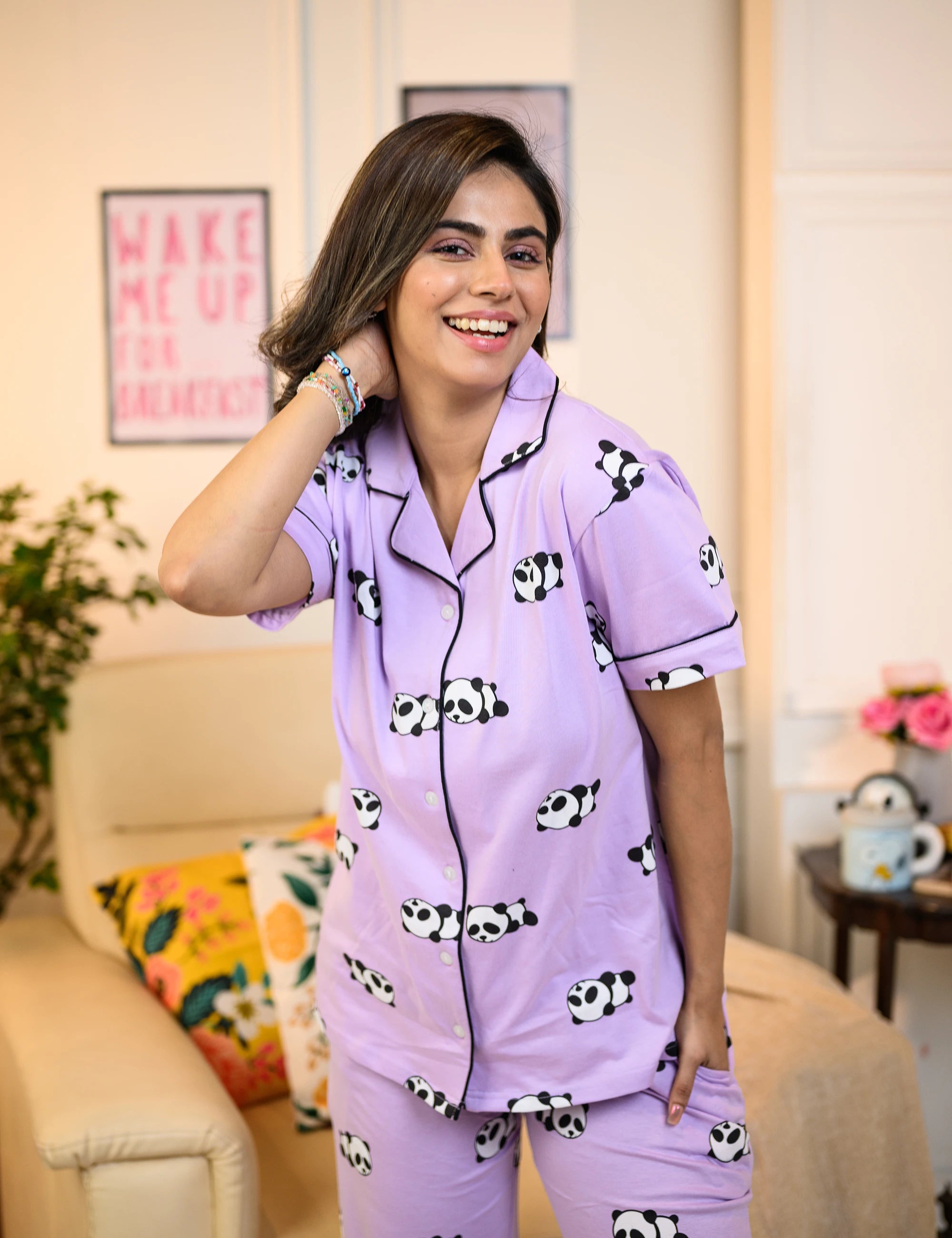 A woman wearing a purple panda print pyjama set, smiling warmly while standing in a cozy, well-decorated room.