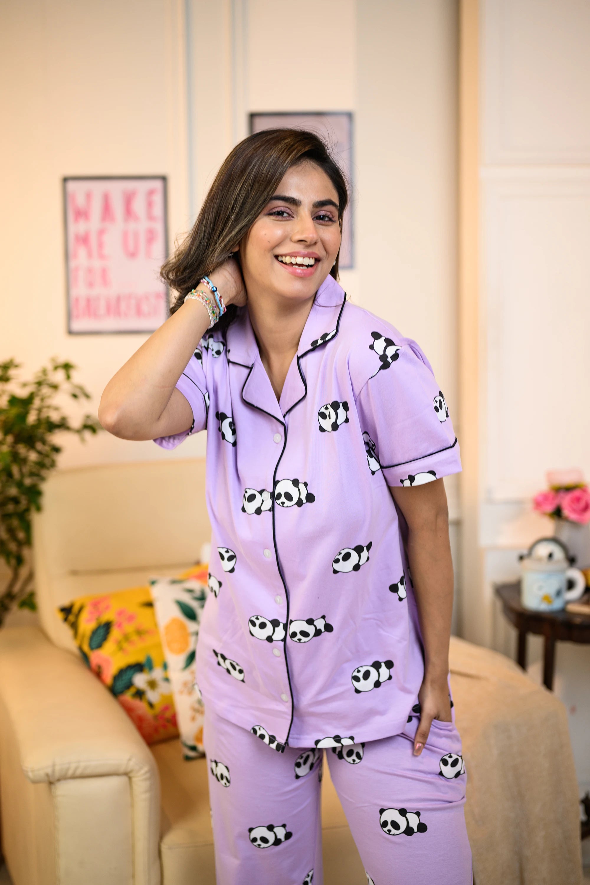 A woman wearing a purple panda print pyjama set, smiling warmly while standing in a cozy, well-decorated room.