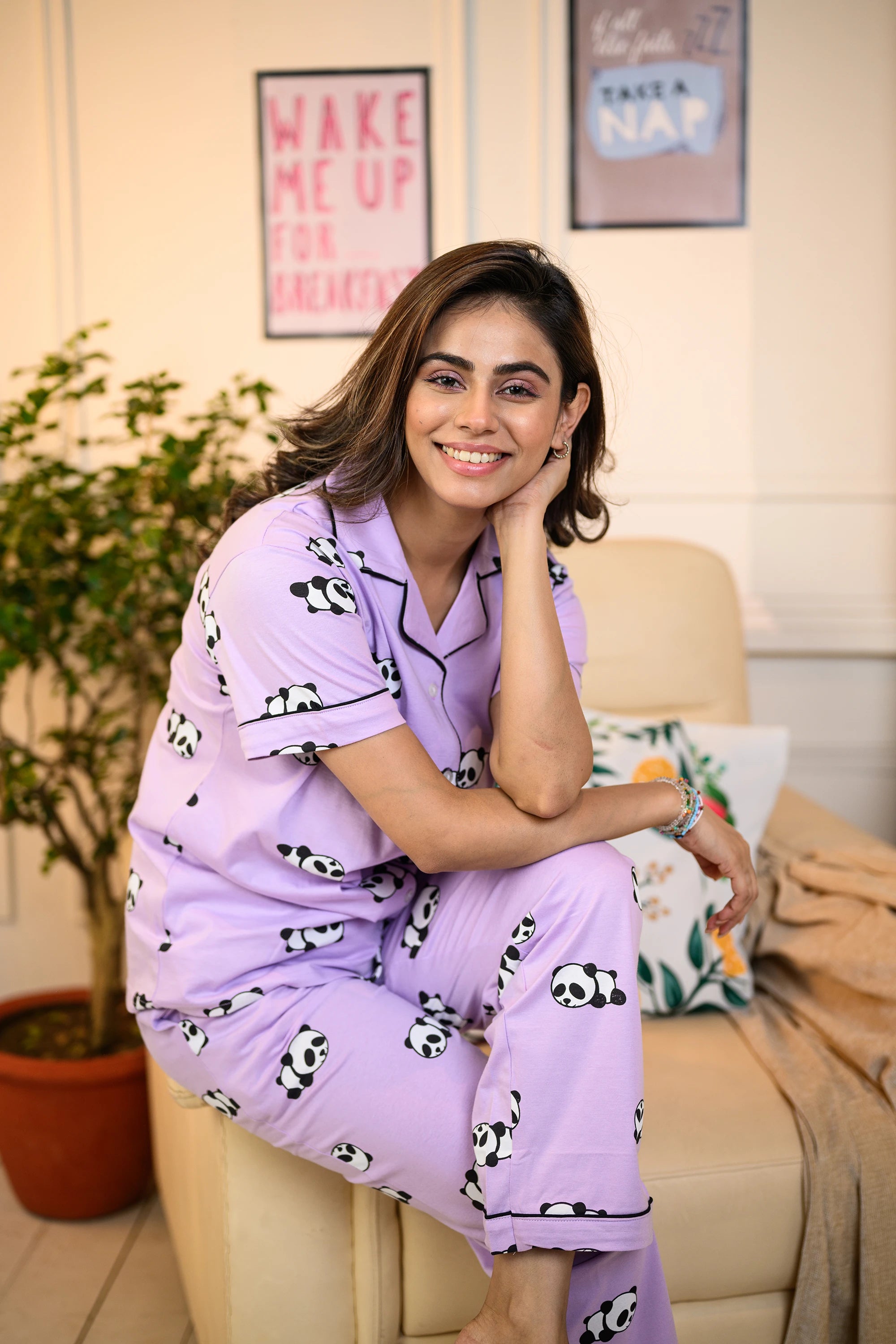 A woman seated on the edge of a sofa chair, wearing a purple panda print pyjama set, smiling warmly in a cozy room