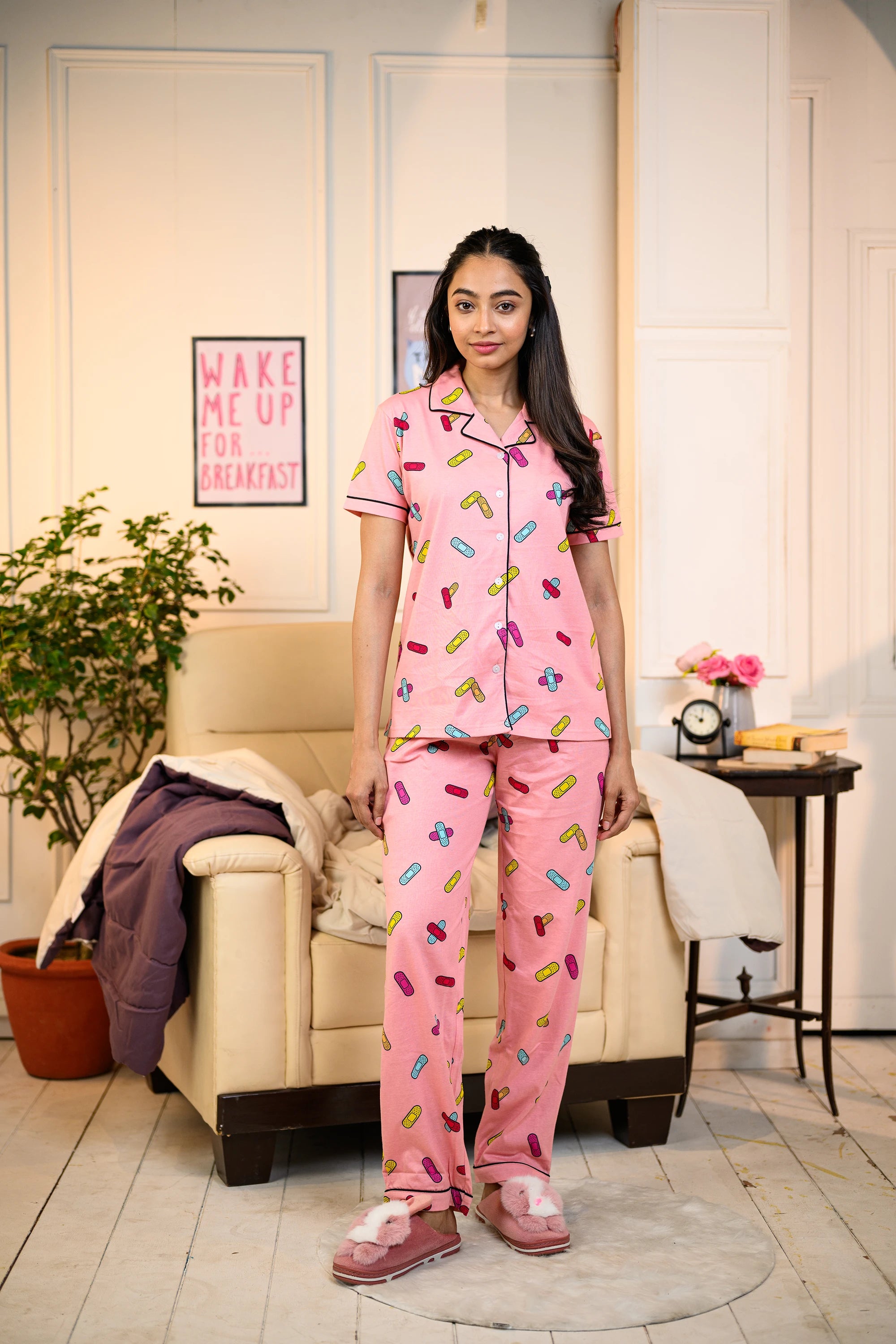 Young woman standing in a cozy living room, dressed in a pink Bandage Print Pyjama Set by NapStory, featuring colorful bandage patterns