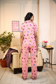 Back view of a young woman wearing a pink Bandage Print Pyjama Set by NapStory, showcasing the vibrant bandage pattern in a cozy living room