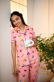 Young woman smiling while holding a coffee mug, dressed in a pink Bandage Print Pyjama Set by NapStory, standing near a bright window with greenery in the background.