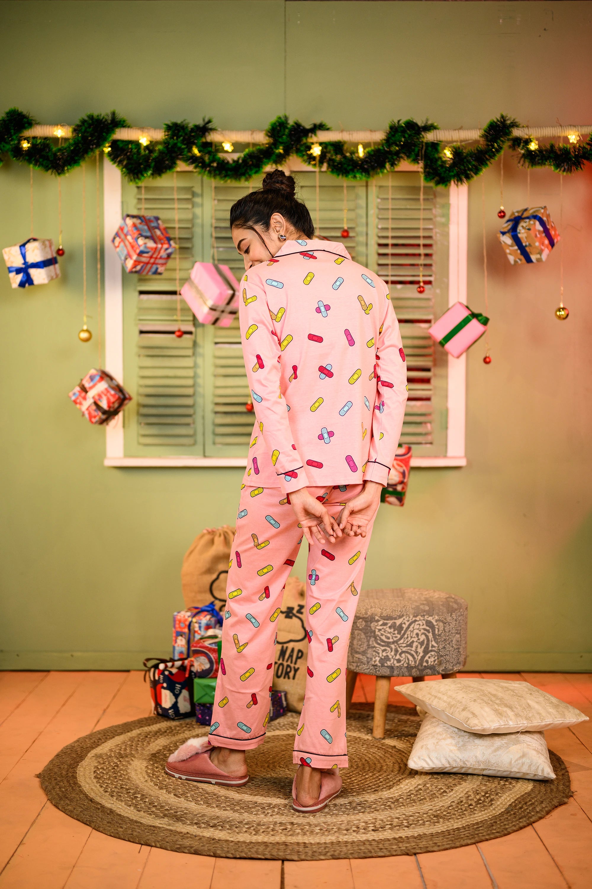 Rear view of the woman standing, wearing the pink bandage print pyjama set, showcasing the back detailing of the outfit, with her hands buckled behind her.