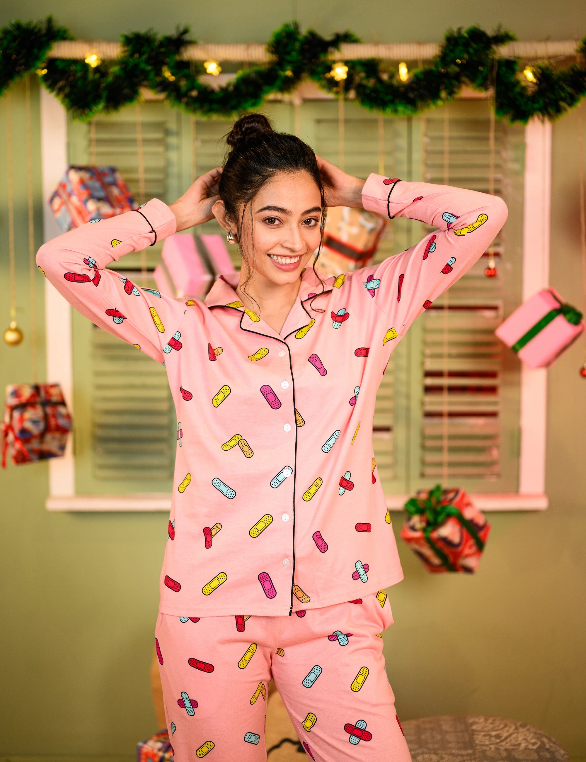 Woman in a relaxed pose, wearing a pink full-sleeved pyjama set with a colorful bandage print, styled with her hair tied back, smiling confidently with her hands behind her head.