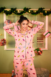 Woman in a relaxed pose, wearing a pink full-sleeved pyjama set with a colorful bandage print, styled with her hair tied back, smiling confidently with her hands behind her head.