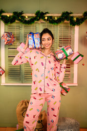 Woman posing playfully while holding two wrapped gift boxes, dressed in the pink bandage print pyjama set, smiling brightly.