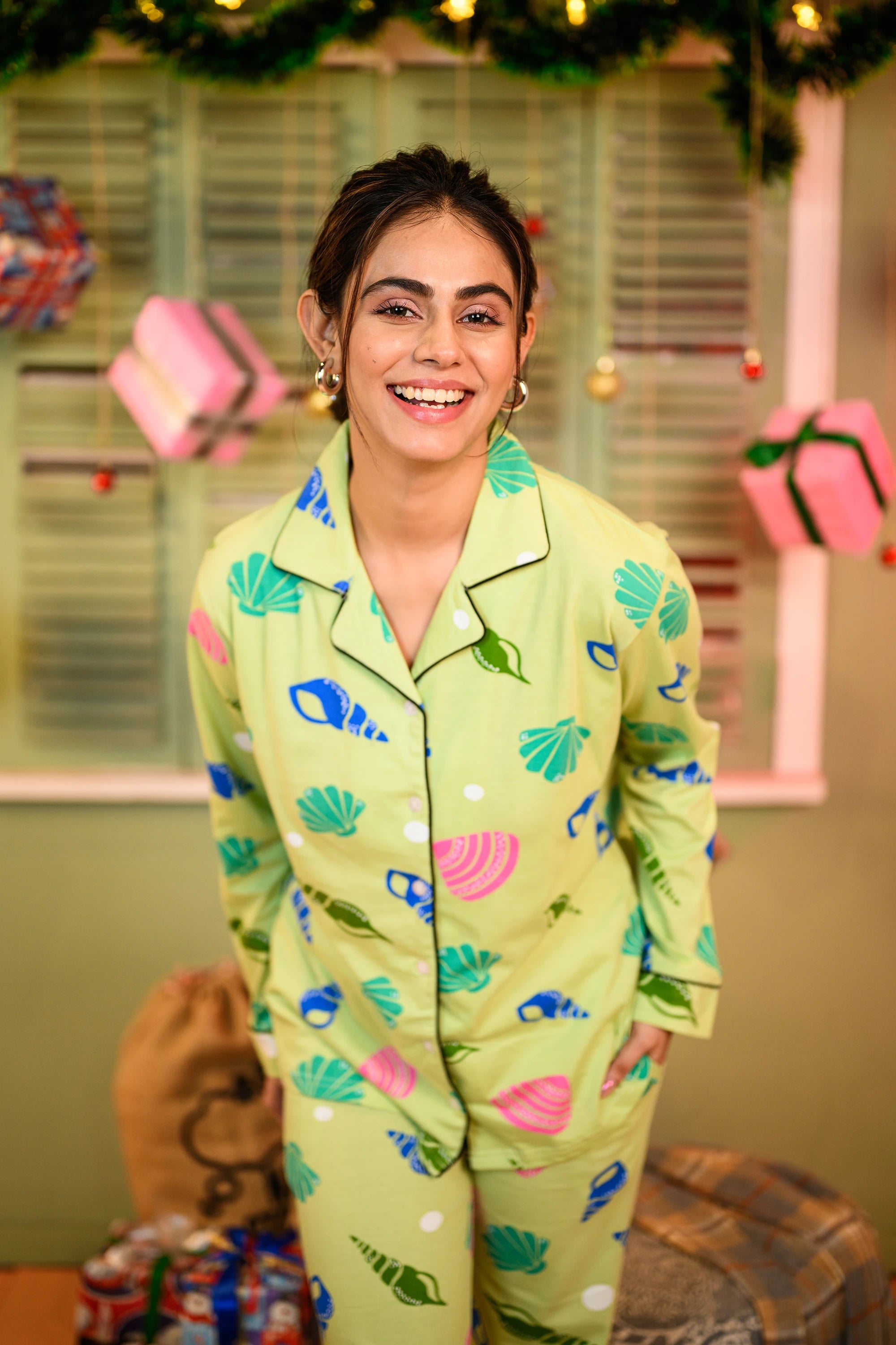 A close-up of a woman standing and leaning slightly forward, wearing a green sea shell print full sleeves pyjama set, smiling brightly.