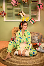 A woman seated on a rug with her legs crossed, wearing a sea shell print full sleeves pyjama set in green, holding a cushion and smiling to her side.