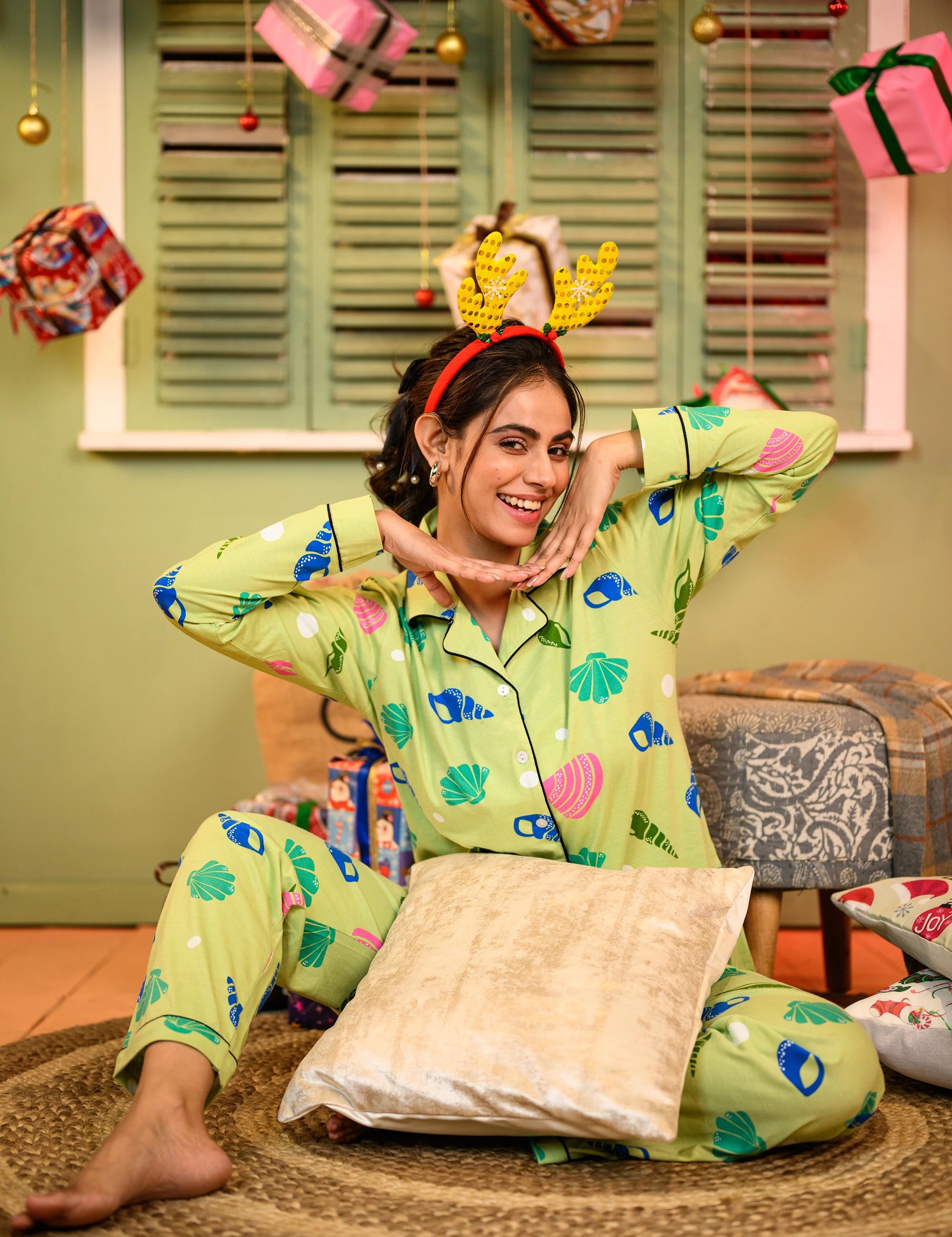 A woman seated cross-legged on a rug, wearing a sea shell print full sleeves pyjama set in green, with her hands framing her face and a playful smile.