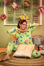 A woman seated cross-legged on a rug, wearing a sea shell print full sleeves pyjama set in green, with her hands framing her face and a playful smile.