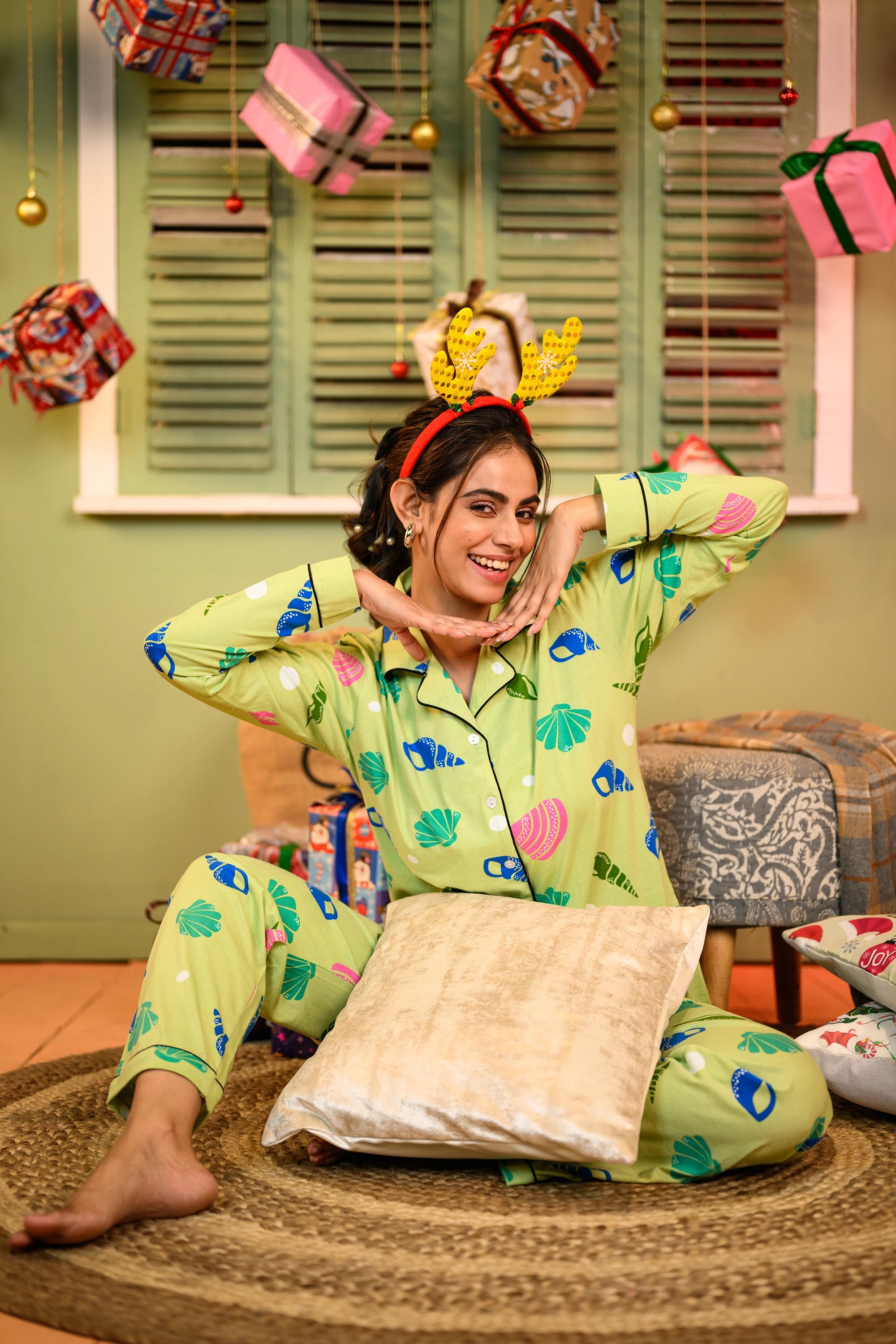 A woman seated cross-legged on a rug, wearing a sea shell print full sleeves pyjama set in green, with her hands framing her face and a playful smile.