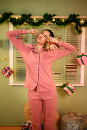 A woman smiling and playfully posing in a Ruddy Pink Full Sleeve Nightsuit Set. She is making a cute face with her hands raised near her eyes, wearing a full-sleeve, buttoned shirt and matching pants, featuring a small embroidered detail on the shirt.